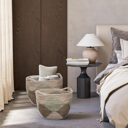 Pair of two nesting handwoven natural brown seagrass storage baskets with integrated handles arranged on a light wood floor in a brightly lit living room.