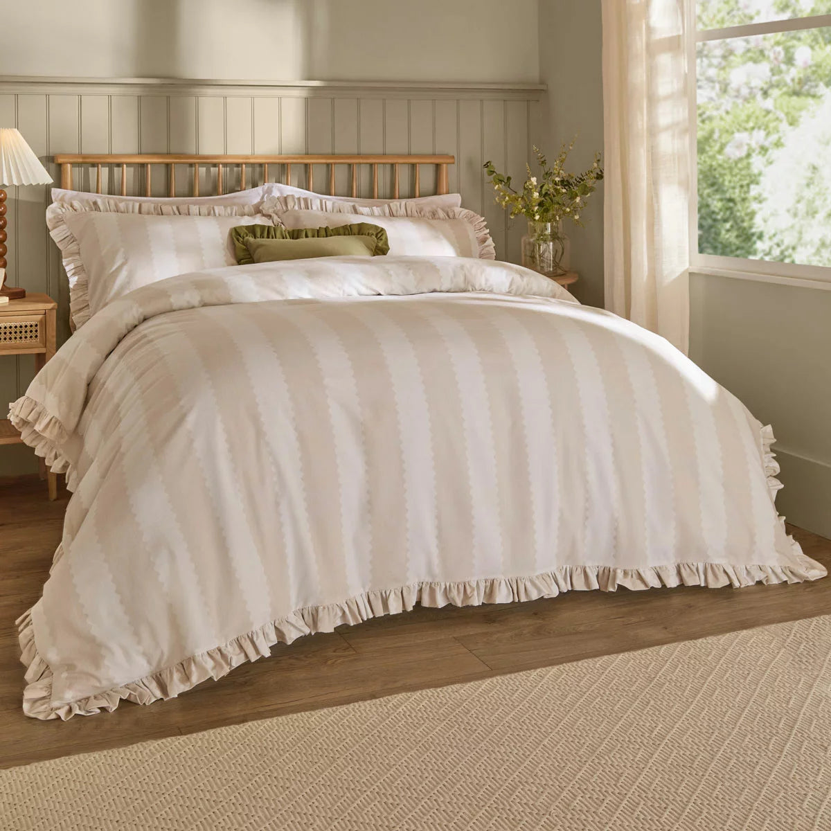 A close-up view of the Wylder Birdie beige stripe duvet cover showing its vintage ruffle trim detail draped over a cozy mattress in a brightly lit bedroom.