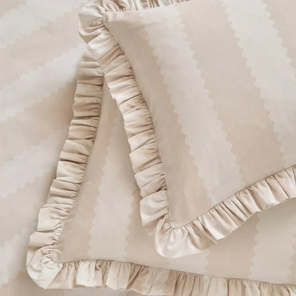 A close-up view of the Wylder Birdie beige stripe duvet cover showing its vintage ruffle trim detail draped over a cozy mattress in a brightly lit bedroom.