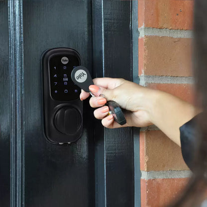 A close-up shot of the matte black Yale Keyless Smart Lock displaying illuminated blue numbers on its digital touch screen, mounted onto a modern dark oak wooden door.