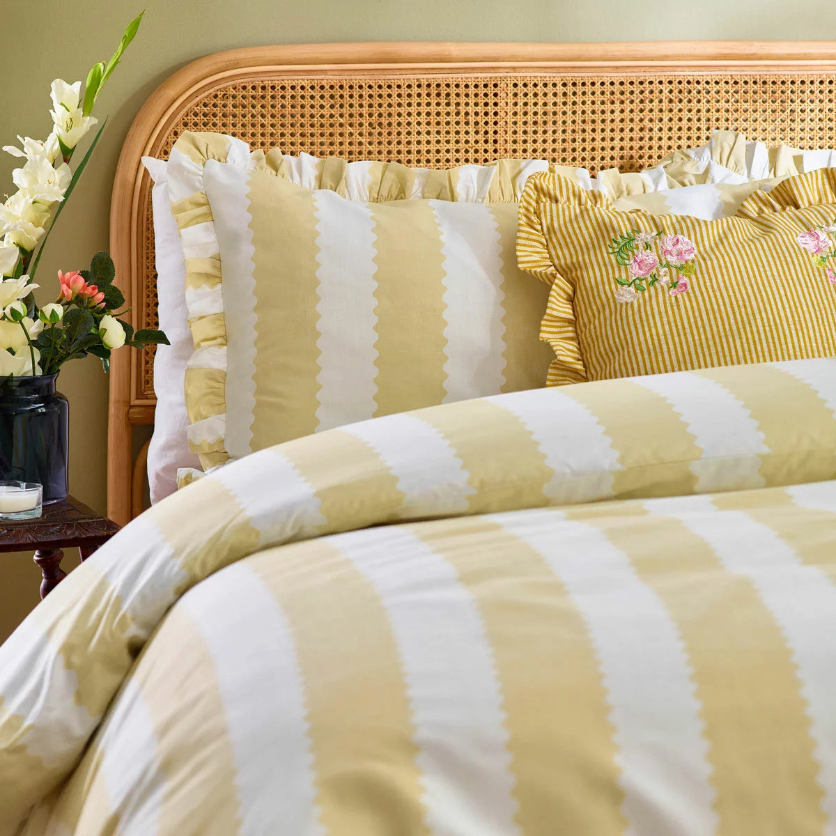 A double bed styled with the yellow-striped Wylder Birdie duvet cover, showcasing the delicate ruffled edges of the pillowcases and cover under warm bedroom lighting.