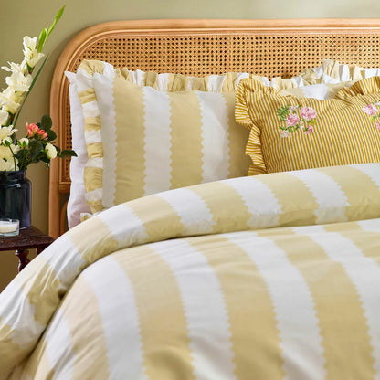 A double bed styled with the yellow-striped Wylder Birdie duvet cover, showcasing the delicate ruffled edges of the pillowcases and cover under warm bedroom lighting.
