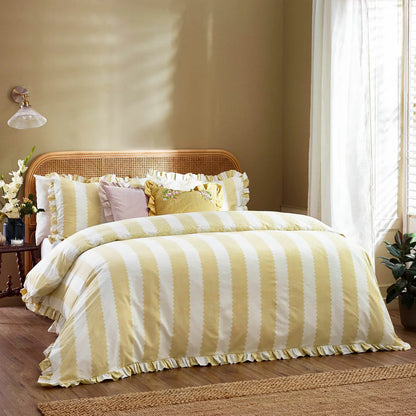 A double bed styled with the yellow-striped Wylder Birdie duvet cover, showcasing the delicate ruffled edges of the pillowcases and cover under warm bedroom lighting.