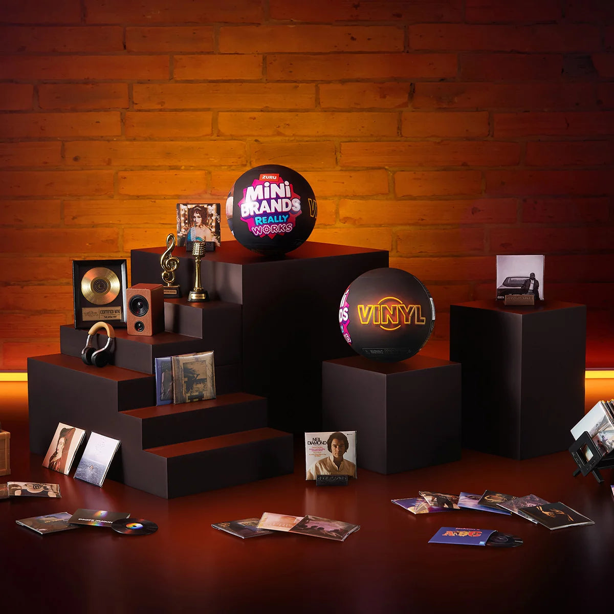 Three colorful ZURU Mini Brands mystery capsules set next to a tiny working black and red mini record player with mini vinyl records spread across a white surface.