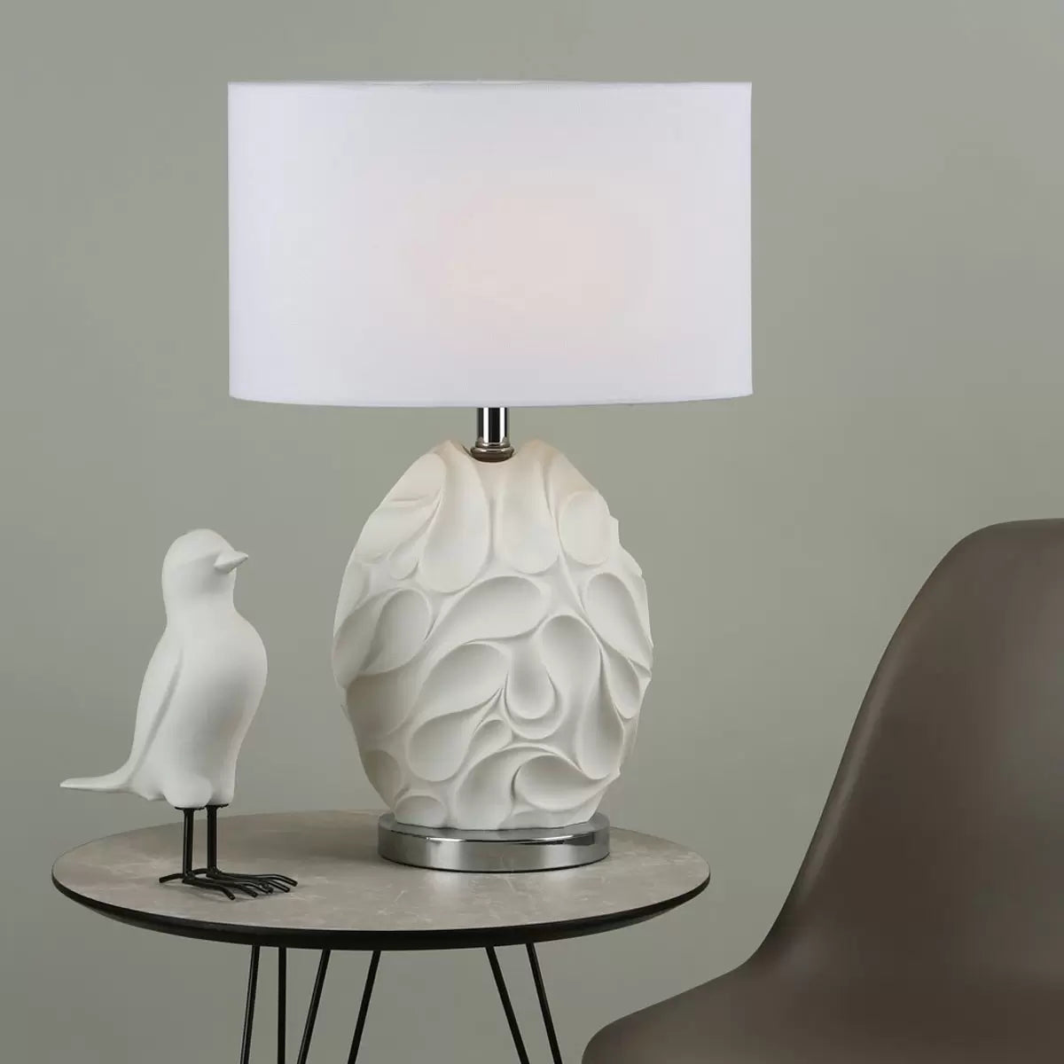 Front view of the matte white textured Zachary oval table lamp with undulating ridges, featuring a polished chrome neck and a white linen oval shade on a minimal concrete sideboard.