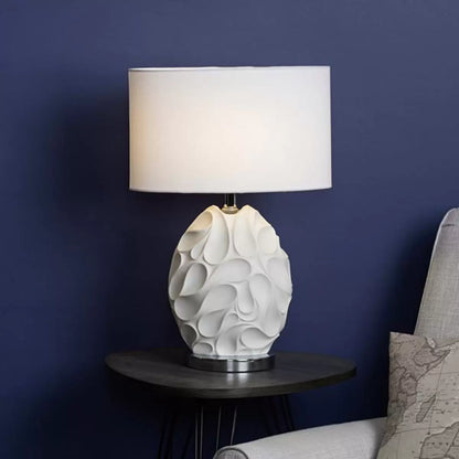 Front view of the matte white textured Zachary oval table lamp with undulating ridges, featuring a polished chrome neck and a white linen oval shade on a minimal concrete sideboard.