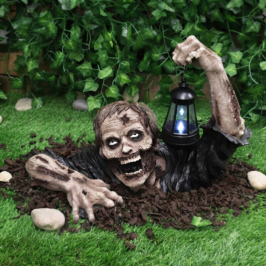 A resin zombie crawling out of a grave holding a glowing LED lantern garden statue, positioned among autumn leaves for a spooky Halloween display.