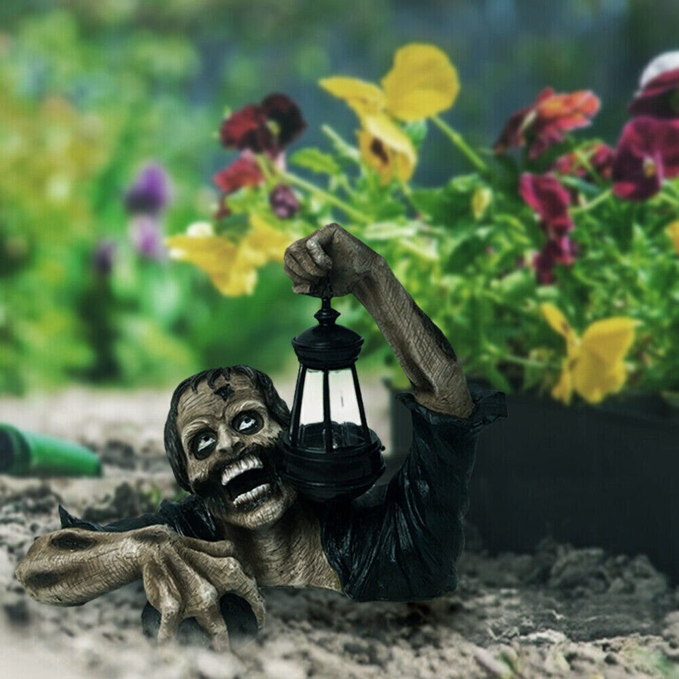 A resin zombie crawling out of a grave holding a glowing LED lantern garden statue, positioned among autumn leaves for a spooky Halloween display.