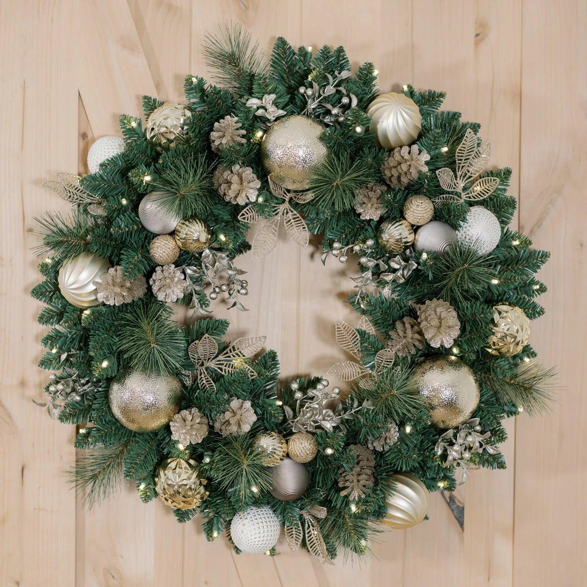 Front view of a 30-inch pre-lit holiday wreath with dense green faux pine needles, natural pinecones, red ornaments, and 50 glowing warm white LEDs displayed on a dark wooden front door.